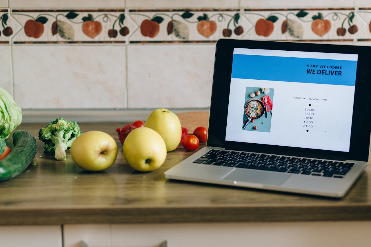 journey Laptop showing a delivery app beside fresh fruits and vegetables on a kitchen counter.